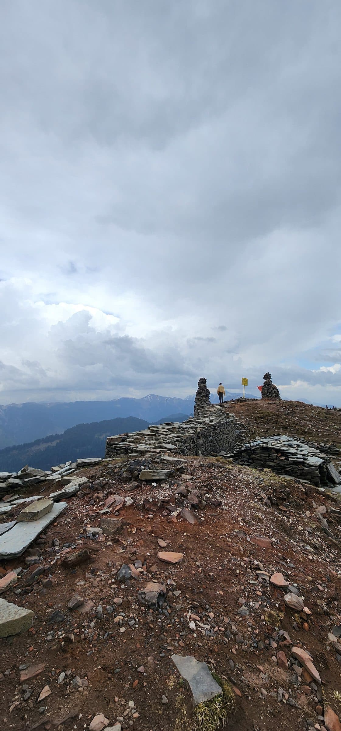 A lone figure stands on a rocky hillside overlooking a scenic valley in Jibhi, Himachal Pradesh, India.