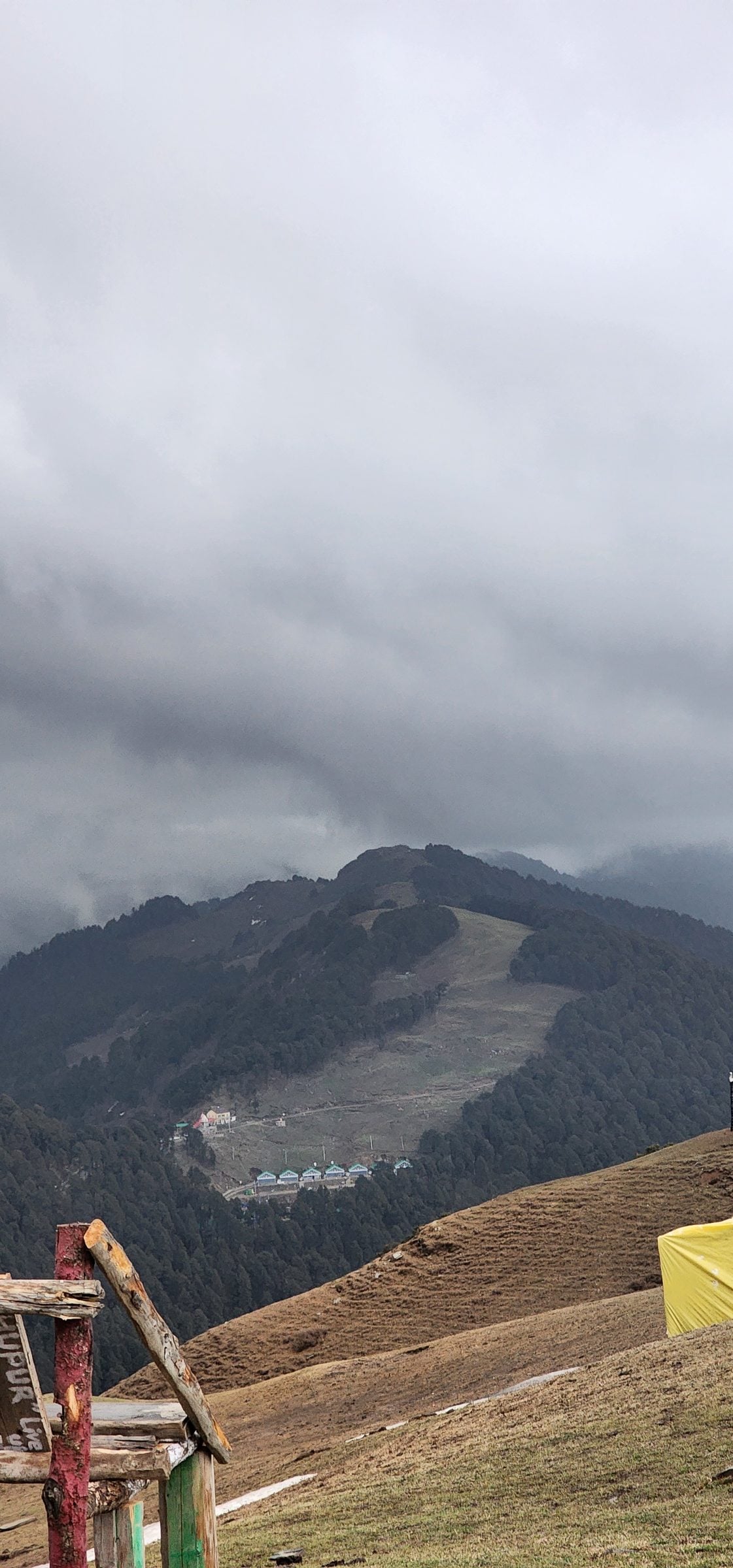 A serene, cloudy hillside view in Shoja, Himachal Pradesh, India, featuring a wooden signpost and distant trees.