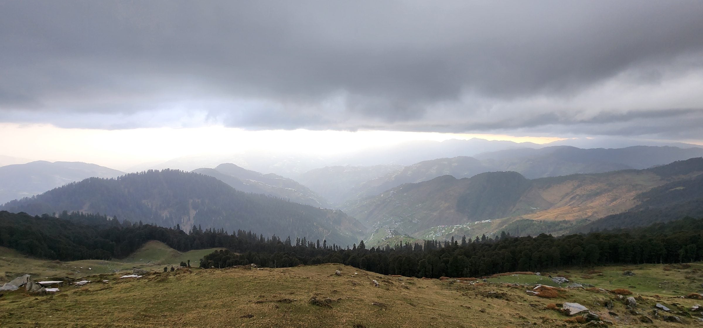 Serene Jibhi Valley: A Himalayan Escape Panoramic view of a lush green valley in Jibhi, Himachal Pradesh, India, featuring scattered trees and a cloudy sky.