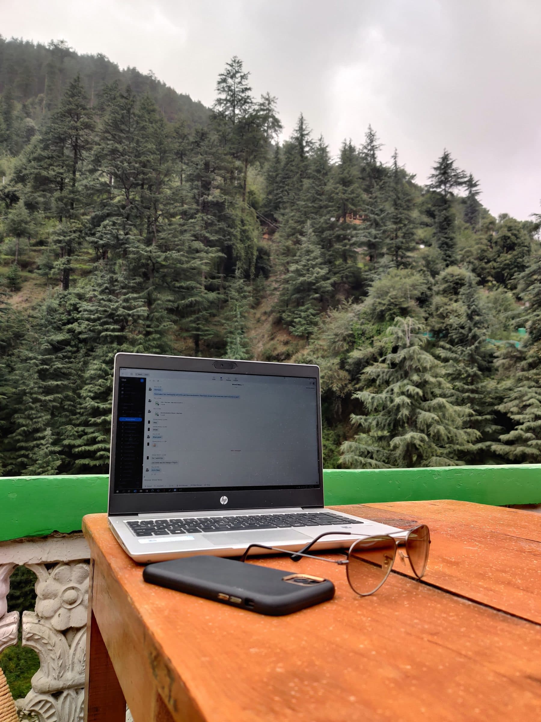 A laptop sits on a wooden table overlooking the lush green hills of Jibhi Valley, Himachal Pradesh.