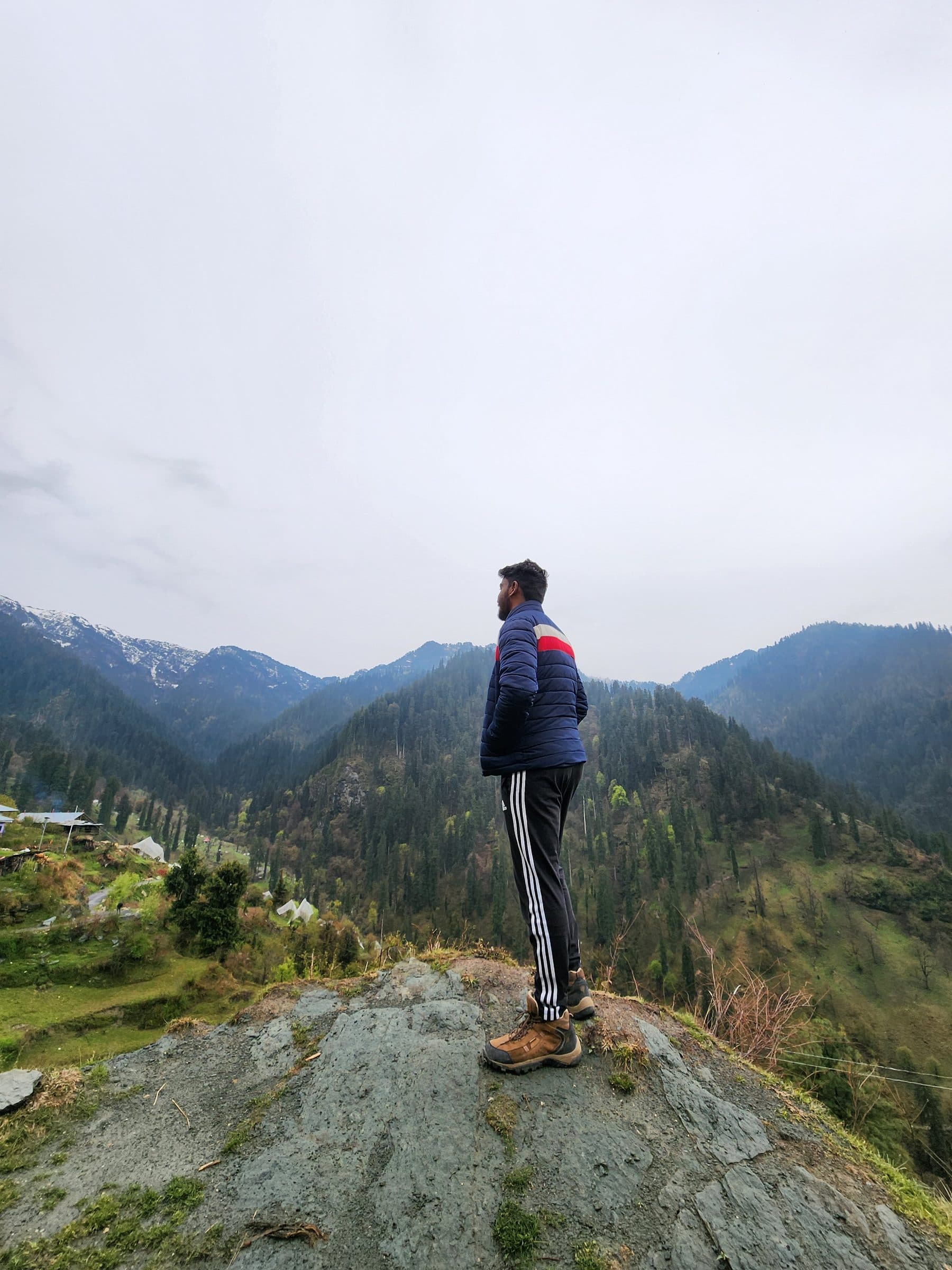 A lone traveler stands on a rocky hillside overlooking a serene valley in Jibhi, Himachal Pradesh, India.
