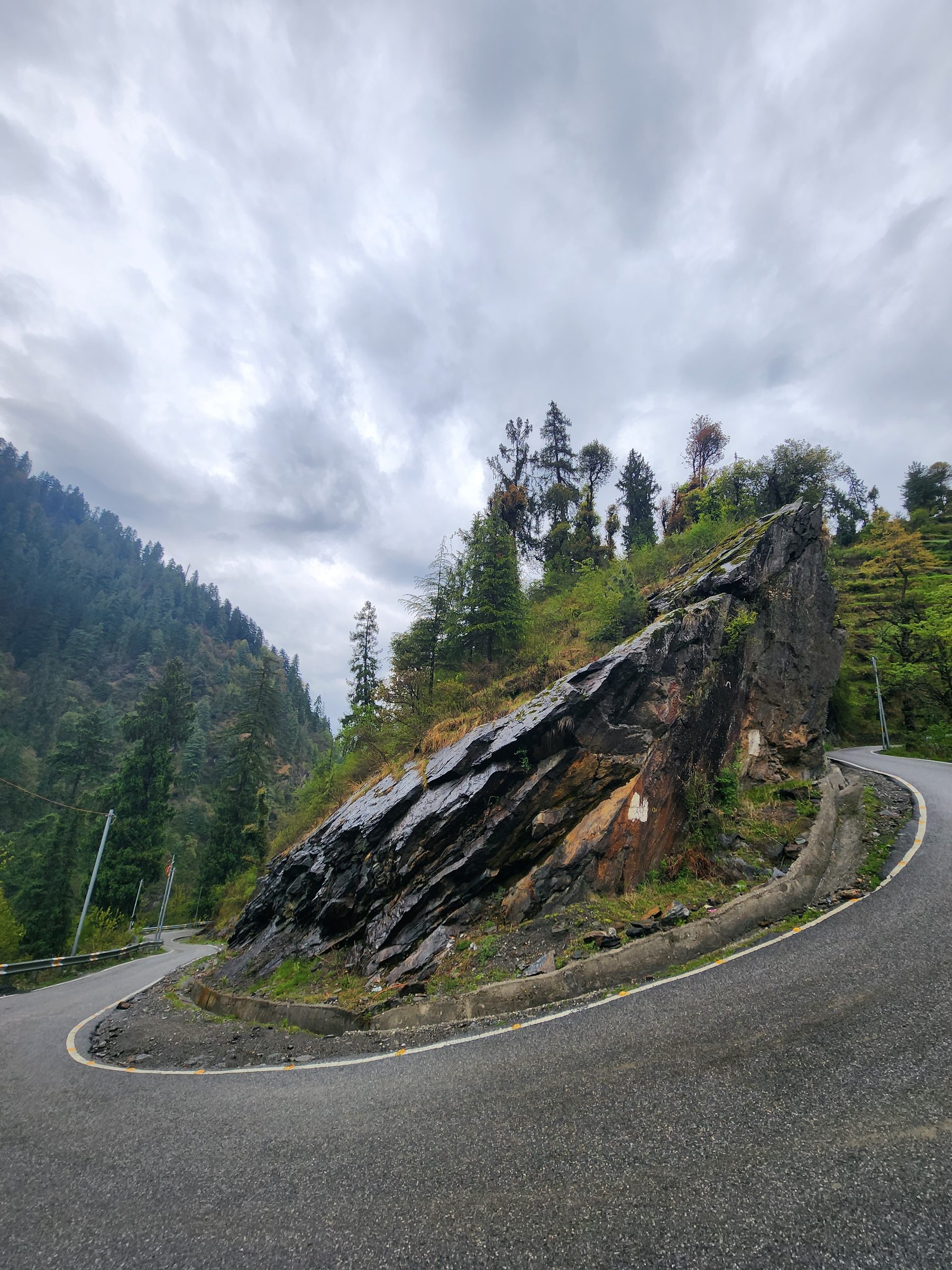 A road curves along a rocky, forested hillside under an overcast sky