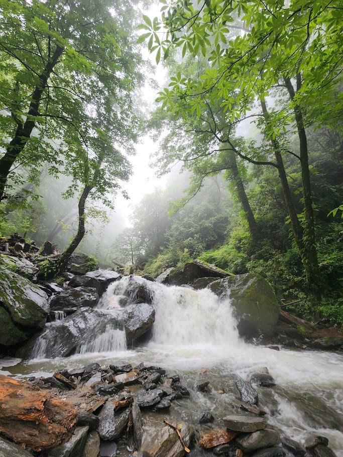 Lush green rocks and trees surrounding a misty flowing waterfall near Ailyak.